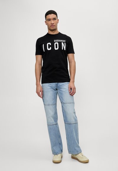 Must musta T-särk, millel on valget värvi "DSQUARED2 ICON" trükk, koos helesiniste teksadega, millel on laienev lõige ja kokkuõmbluse detailid.