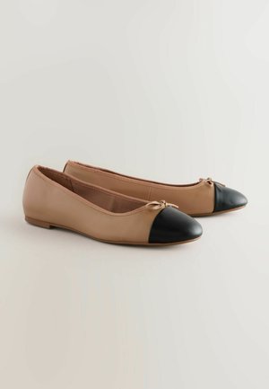 Des ballerines en cuir beige lisse avec un bout rond noir. Elles présentent un petit nœud à l'avant et un design à profil bas.