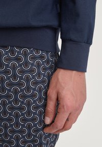 Marineblauwe sweatshirt met geribbelde boorden, gecombineerd met een patroonbroek met bogen in wit, bruin en lichtblauw op een donkerblauwe achtergrond.
