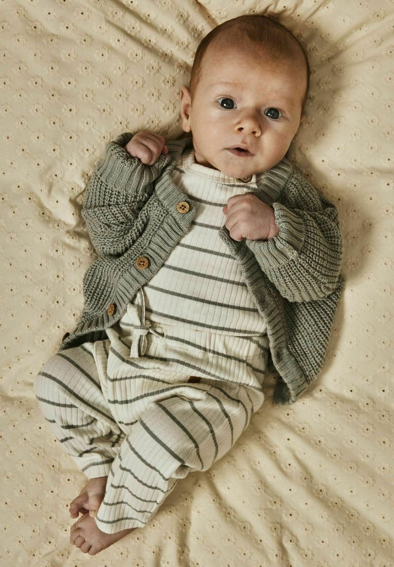 Cardigan verde lavorato a maglia con bottoni in legno, sopra un body a righe chiaro e pantaloni, poggiato su uno sfondo di tessuto beige texture.