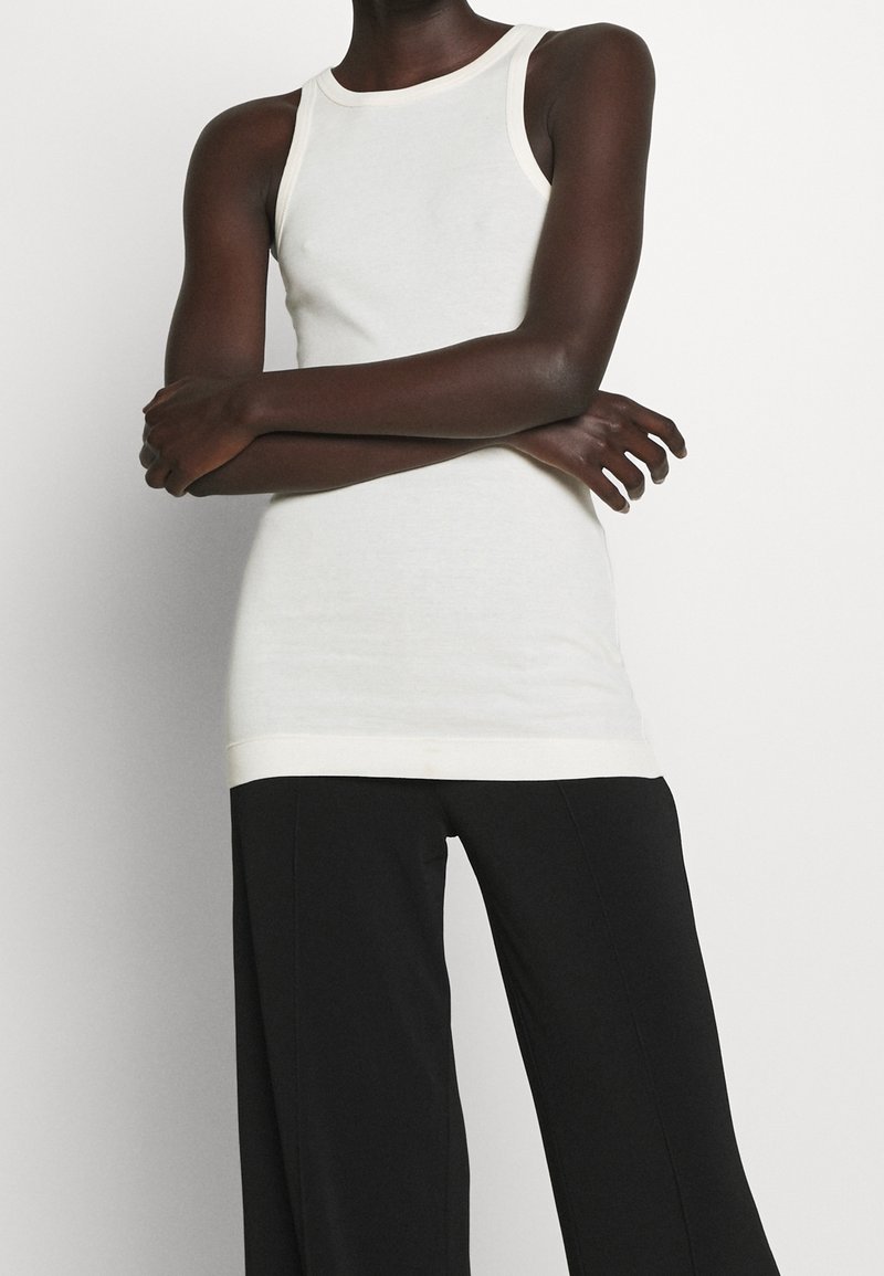 Débardeur blanc sans manches au design ajusté avec un ourlet beige contrastant, associé à un pantalon large noir avec une texture lisse.