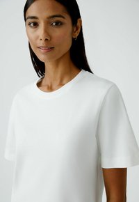 Weißes T-Shirt mit kurzen Ärmeln aus weichem Material, mit einem Rundhalsausschnitt und dezent geprägtem Logo auf der Brust. Glatte Textur.