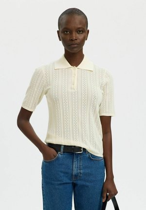 Femme aux cheveux courts portant un polo en maille crème, un jean bleu avec une ceinture noire, main dans la poche, tenant une sangle noire sur un fond uni.