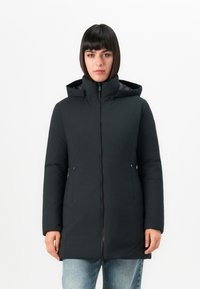 Frau mit glattem, dunklem Haar, die einen schwarzen Winterschafts mit Kapuze trägt und zugezippt ist, steht vor einem schlichten weißen Hintergrund.