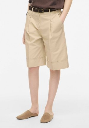 Personne portant un short beige jusqu'aux genoux avec une ceinture marron et des chaussures marron sans lacets, debout les mains dans les poches.