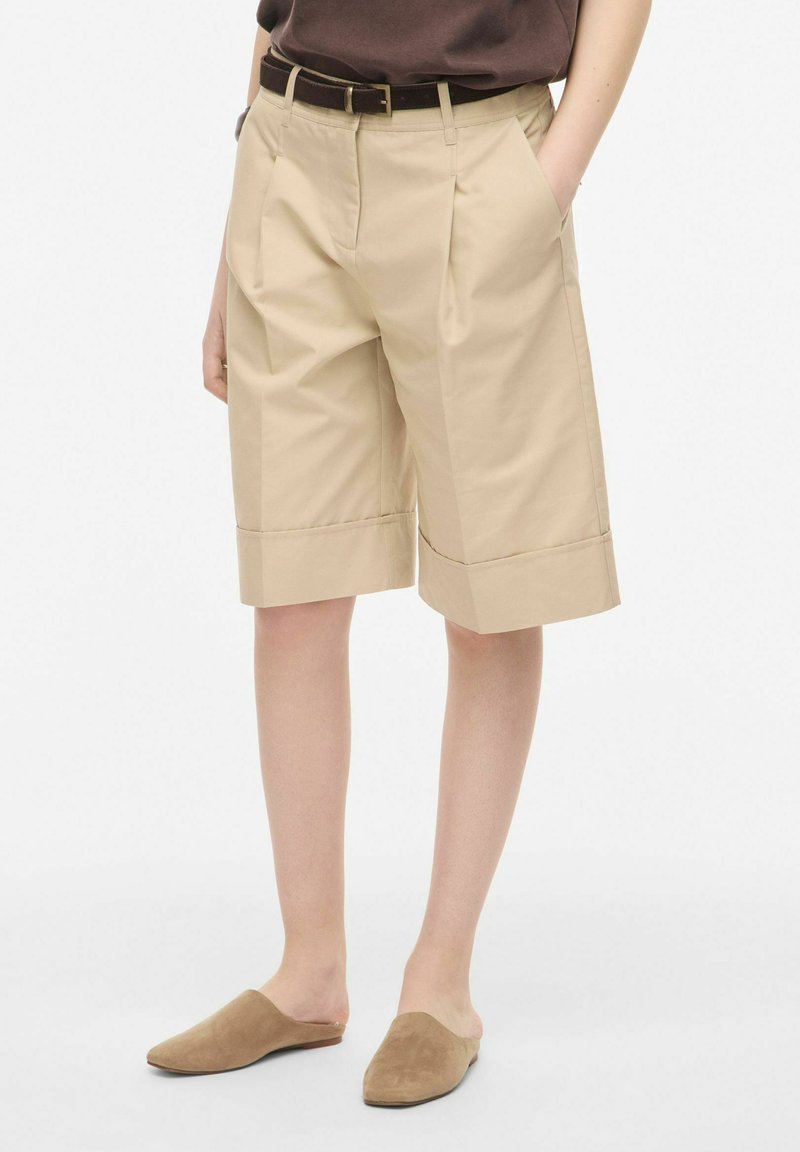 Personne portant un short beige jusqu'aux genoux avec une ceinture marron et des chaussures marron sans lacets, debout les mains dans les poches.