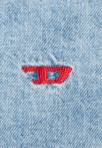 Denim fabric featuring a red embroidered logo, consisting of a stylized "D" integrated into a geometric shape. Light blue background.
