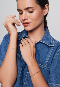 Na nadgarstkach modelki znajdują się dwa delikatne bransoletki. Jedna z nich zdobiona jest kolorowymi koralikami, a druga ma czarne i srebrne koraliki. Modelka ma na sobie dżinsową koszulę.