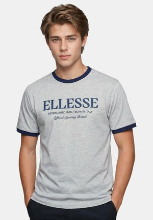Jeune homme portant un T-shirt gris clair avec un col et des manches bleu marine, logo "ELLESSE" et texte centrés sur la poitrine, mains dans les poches.