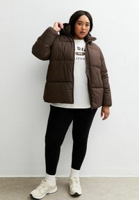 Brązowa puchowa kurtka z kapturem, z zamkiem błyskawicznym z przodu i pikowanym wzorem. Zestawiona z białą koszulką z grafiką, czarnymi legginsami i jasnymi sneakersami.