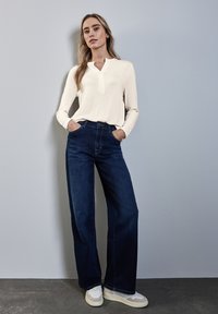 Cremfarbendes Langarmshirt mit Henley-Ausschnitt, kombiniert mit dunkelblauen weit geschnittenen Jeans und hellen Sportschuhen. Glatter Stoff und strukturierte Passform.