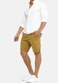 Shorts en coton marron avec des poignets roulés et des poches latérales, associés à une chemise en lin blanche à boutons et à des baskets blanches.