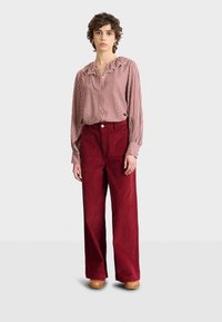 Pantaloni larghi in corduroy rosso con tasche frontali, abbinati a una blusa a scacchi con colletto arricciato. Tacchi a piattaforma in legno completano il look.