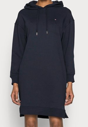 Kvinde iført en knælængde marineblå hættetrøjekjole med lange ærmer og et lille logo på brystet.