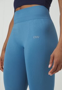 Leggings sportivi blu realizzati in tessuto elastico, con una vita alta a costine e un logo discreto sulla coscia. Dettaglio con pannello laterale testurizzato.