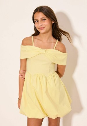 Teenager-Mädchen mit langen braunen Haaren, das ein schulterfreies gelbes Kleid mit Schleifendetail trägt, steht vor einem hellen Hintergrund.