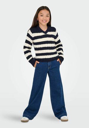 Jong meisje dat glimlacht, gekleed in een zwart-wit gestreepte trui, wijde blauwe jeans en witte sneakers, staande met haar handen in haar zakken.