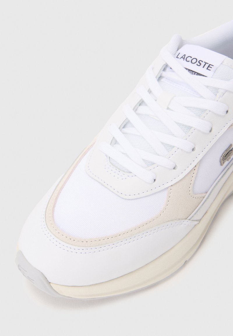 Scarpa da ginnastica bianca realizzata con materiali misti, con una tomaia bianca liscia, dettagli in suède beige, lacci e una suola con texture. Logo Lacoste visibile.