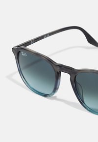 Ochelari de soare Ray-Ban cu un cadru translucid de culoare gri-negru și lentile teal cu degradé. Prezintă o formă rotundă și un logo pe bratul ochelarilor.