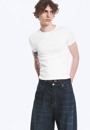 Jeune homme aux cheveux bouclés portant un t-shirt blanc uni et un jean large foncé, les mains dans le dos, regardant sur le côté.