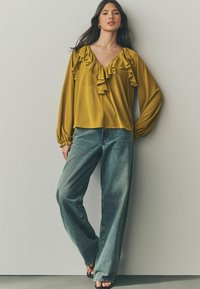 Senapsgul blus med volangkrage och långa ärmar, ihopparad med ljusblå vida jeans och svarta klacksandaler.