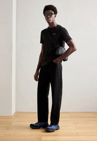 T-shirt noir à manches courtes avec petit logo, pantalon large noir et baskets bleu-noir. Sac à la main, debout sur un sol en bois.