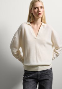 Pull léger en crème surdimensionné avec un col en V et des poignets côtelés. Texture tricotée, coupe décontractée, associé à un jean gris foncé.
