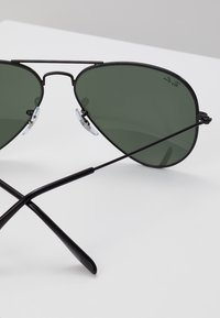 Ochelari de soare aviator negri cu lentile verzi, cadru metalic și pernuțe transparente pentru nas; bratele se împletesc sub cadru, evidențiind un design elegant.