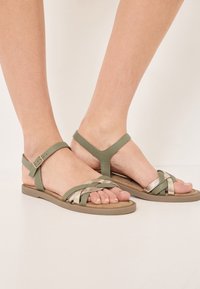 Sandali in pelle verde con cinturini intrecciati in argento chiaro, suola piatta, fascetta alla caviglia regolabile e design minimalista a T.