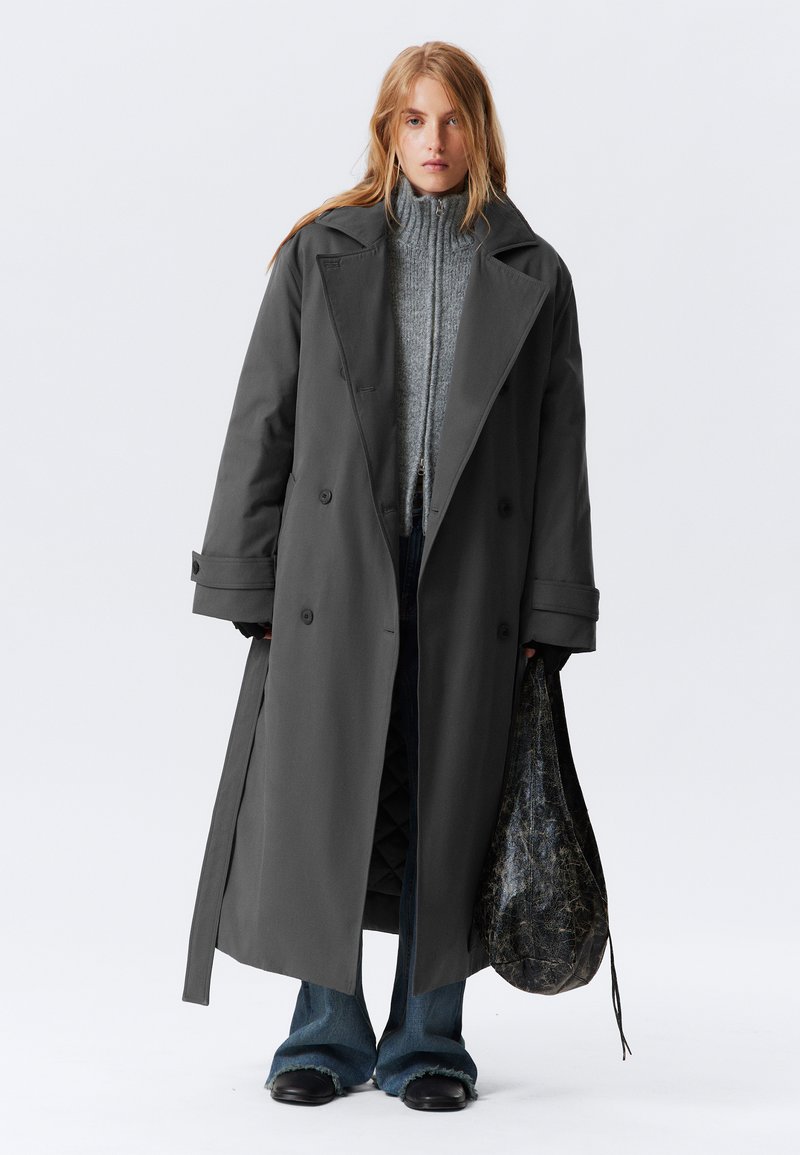 Lungo cappotto grigio con design doppiopetto. Indossato sopra un maglione grigio lavorato a maglia, abbinato a jeans a zampa d'elefante e scarpe in pelle nera. Tiene una borsa con motivo.