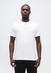 Άνδρας όρθιος, φοράει ένα απλό λευκό t-shirt Calvin Klein και μαύρο παντελόνι, με φόντο ανοιχτό γκρι.