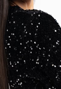 Gros plan sur l'épaule d'une personne aux longs cheveux portant un vêtement noir couvert de paillettes, sur fond blanc uni.