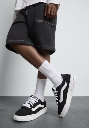 Schwarz-weiße Sneakers aus Wildleder und Textil mit einer dicken weißen Sohle, Schnürsenkeln und charakteristischem Logo. Kombiniert mit weißen Rippensocken.