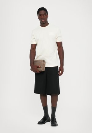 Mann iført kremfarget kortermet t-skjorte, svarte kne-lange shorts, mørke sokker, svarte loafers, holder en taupe clutch veske, står på hvit bakgrunn.