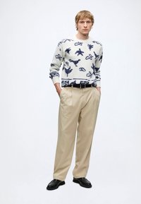 Jeune homme portant un pull blanc avec des motifs animaux bleu marine, pantalon plissé beige, ceinture noire et chaussures noires, debout les mains dans les poches.