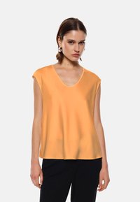 Top arancione senza maniche con scollo a V profondo, texture liscia, vestibilità ampia e maniche corte strutturate, abbinato a pantaloni scuri.