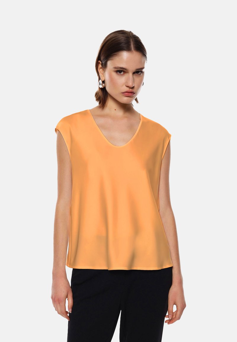 Top arancione senza maniche con scollo a V profondo, texture liscia, vestibilità ampia e maniche corte strutturate, abbinato a pantaloni scuri.