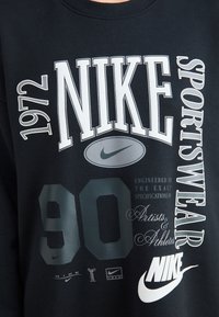 Černá mikina s kombinací bílého a šedého textu, obsahujícího "NIKE," "1972" a "90," s logem Nike swoosh a různými grafickými detaily.