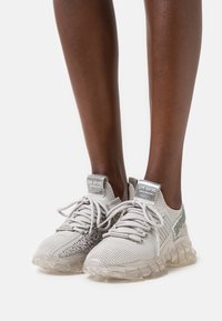 Helle graue Sportschuhe mit einem Obermaterial aus gestricktem Mesh, strukturierten Akzenten, transparentem Gummisohle und reflektierenden Details an Zunge und Seiten.