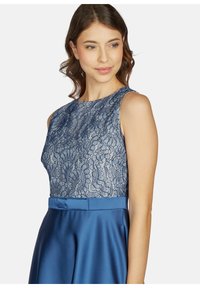 Ein ärmelloses Kleid mit einem marineblauen Spitzenoberteil und einem glatten, satinierten blauen Rock. Das Kleid hat eine taillierte Taille mit einem Schleifenakzent.