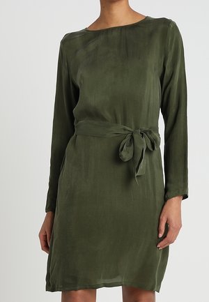 Žena má na sobě olivově zelené šaty délky po kolena s dlouhými rukávy, kulatým výstřihem a látkovým pásem uvázaným v pase.