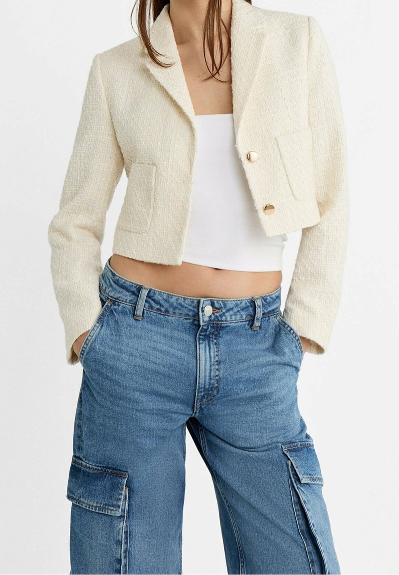 Femme portant un blazer court crème texturé avec des boutons dorés, un haut tube blanc, et un jean cargo bleu ample les mains dans les poches.