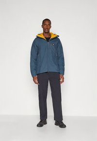 Mėlyna izoliuota striukė su geltona pamušalu, kišenėmis su užtrauktukais ir logotipu krūtinės srityje, derinama su juodomis kelnėmis ir tamsiais batais.