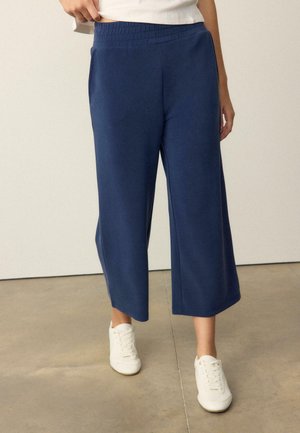 Personne portant un pantalon large bleu marine raccourci et des baskets blanches, debout sur un sol en béton poli devant un mur uni.