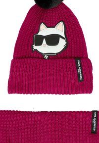 Beanie in magenta brillante con pompon nero, con una grafica di un gatto bianco che indossa occhiali da sole e un'etichetta con logo sul polsino.