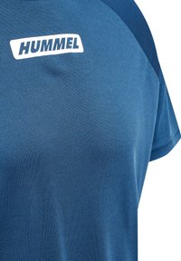 Blå sports shirt med tekstureret stof og hvidt HUMMEL-logo på øverste bryst, korte ærmer og mørkeblå skulderpaneler.