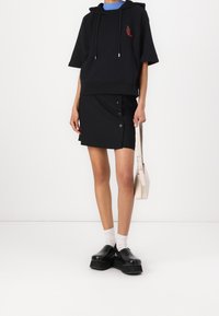 Černá oversized mikina s výšivkou červené papričky, černá mini sukně s bočními knoflíky, bílé ponožky a masivní černé platformové boty.