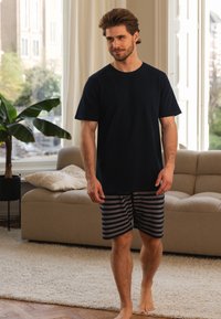 Marineblauwe katoenen T-shirt met korte mouwen, gecombineerd met grijs gestreepte shorts. Lichte huid en haar, op blote voeten, in een heldere kamer met een bank.
