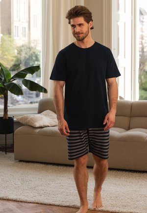 Marineblauwe katoenen T-shirt met korte mouwen, gecombineerd met grijs gestreepte shorts. Lichte huid en haar, op blote voeten, in een heldere kamer met een bank.