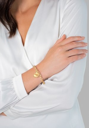 Gouden armband met hart bedel, met een schakeldesign. Draagt op een pols, vergezeld van een witte, iets gestructureerde blouse.